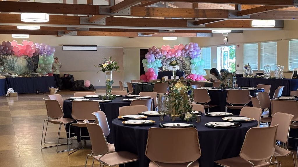 Multipurpose room set up with round tables, mauve chairs, rented linens and tableware, there are balloon arches in the background.